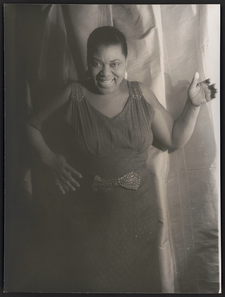 Carl Van Vechten's 1936 photograph of Bessie Smith standing with her right arm raised. She is smiling with her head slightly tilted down.
