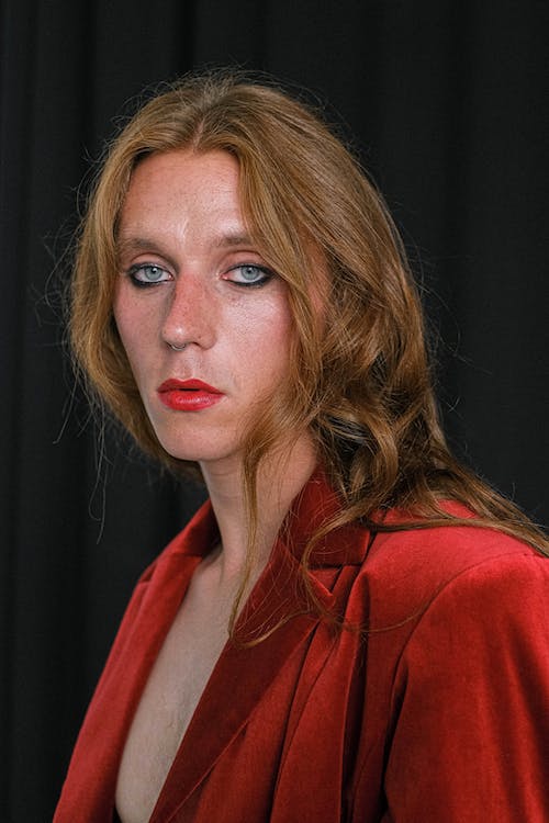 Serious young androgynous male model with makeup and long ginger hair in red blazer standing against black background and looking at camera thoughtfully