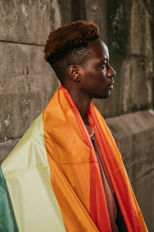 Contemplative young African American homosexual male with dyed hair standing on street with colorful LGBT flag on shoulders and looking away